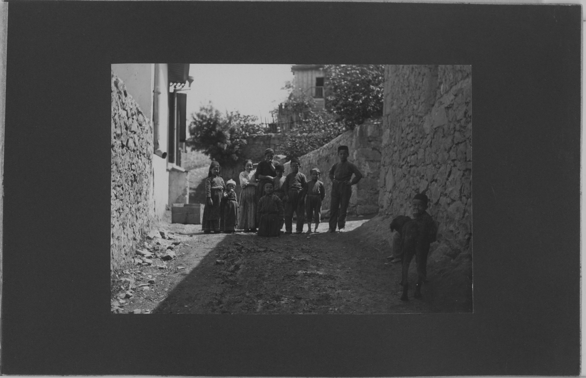 'Grupp barn, ca 10 barn, samlade i en gränd tillsammans med en hund. Med stenmursväggar på sidorna. ::  :: Se serie med fotonr. 5270:1-15. Serie fotonr 5269-5292, Stuxbergs resa till Kaukasien, 1897.'