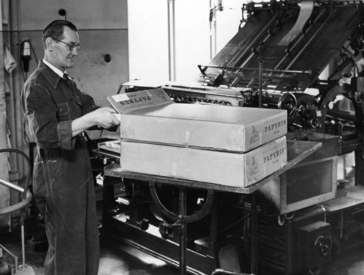 Tryckerilager med införpackningar av Wellpappkartonger, användning i tegel..? på Papyrus, den 15/11-1958.
En man arbetar vid en maskin.