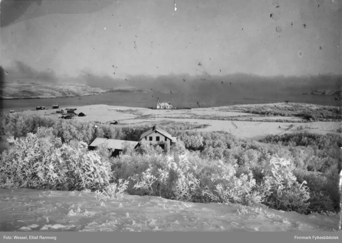 Bildet tatt fra Kirkenes før anlegget kom. Føran i bildet distriktslegeboligen, bak den klokker Anders Tokles gård og lengts bak kirke. Landskapet er dekt av snø.
