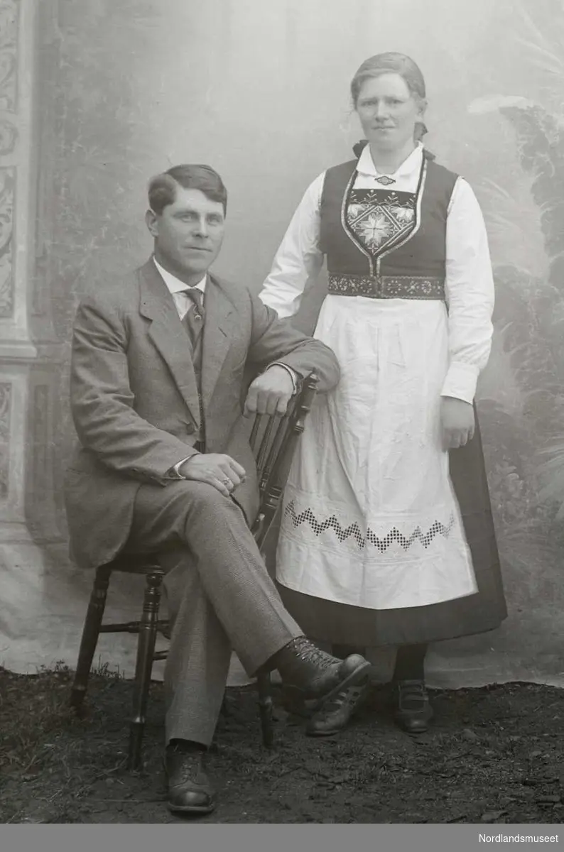 Portrett. Par i 30-40-åra. Mannen sitter, iført dress. Kvinnen står ved siden av i bunad. Disse to finner vi igjen i NM 8243:648.