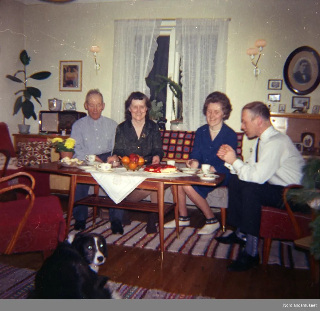 Familieportrett? i interiør.
To menn og to kvinner ved kaffebordet.
I forgrunnen liggende hund.