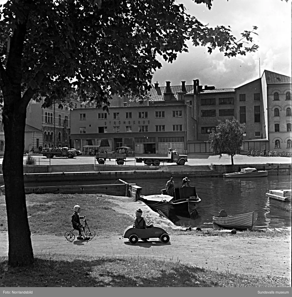 En båt går in vid en liten strand som fanns utgrävd på Selångersåns norra sida mitt emot Strömbergs Färg i kvarteret Lyckan. I förgrunden två lekande barn.