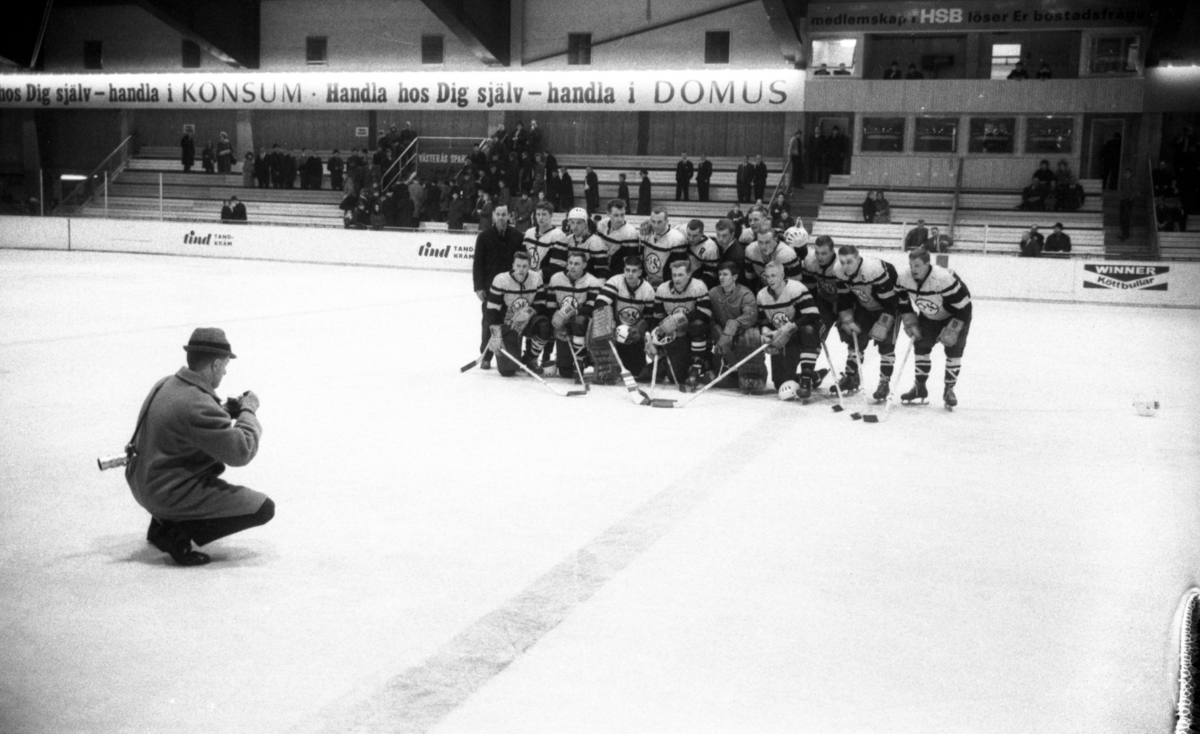 Ishockey klar 31 januari 1966

Ett ishockeylag i idrottskläder samt med ishockeyklubbor i händerna står uppställda på isen för att bli fotograferade. En fotograf med kamera sitter framför dem på isen. Publik står på läktaren och betraktar det hela.