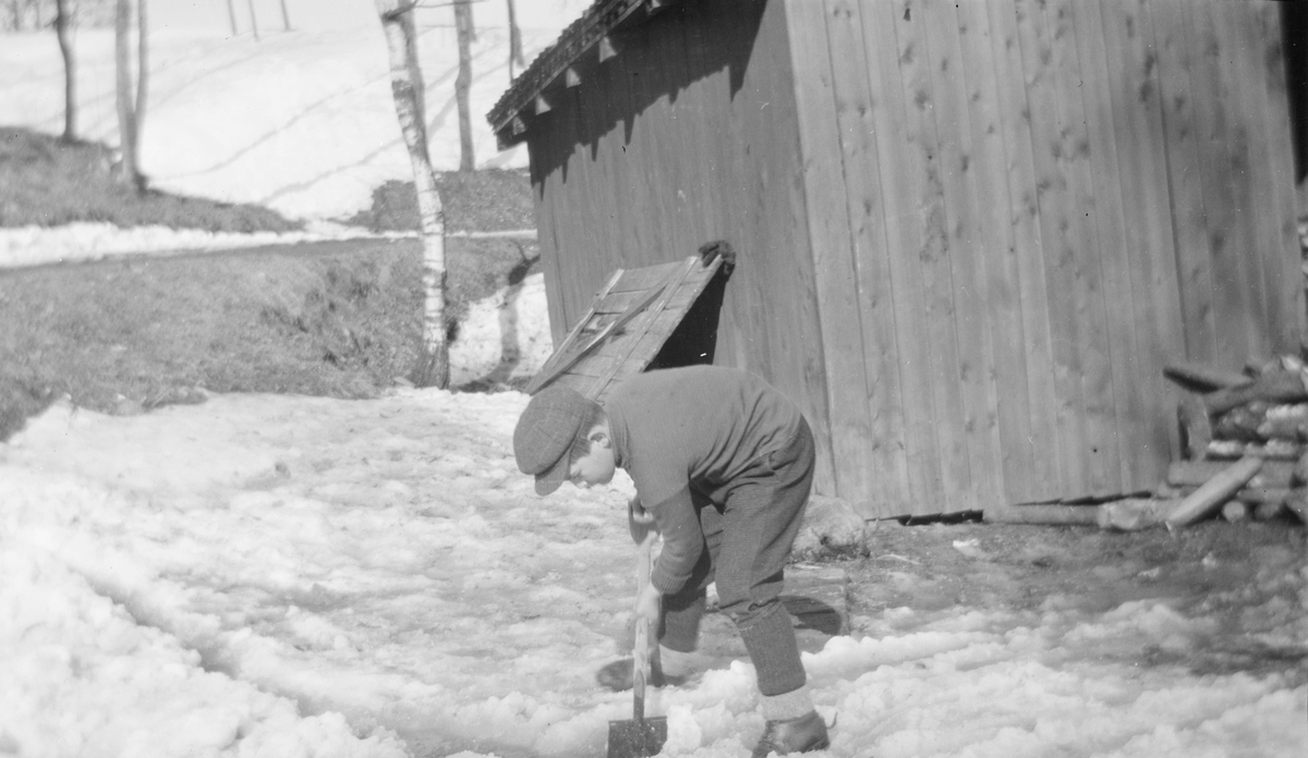 En gutt, trolig Haaken Christian Mathiesen f. 1896 måker våt snø med en spade. Uthus i bakgrunnen.