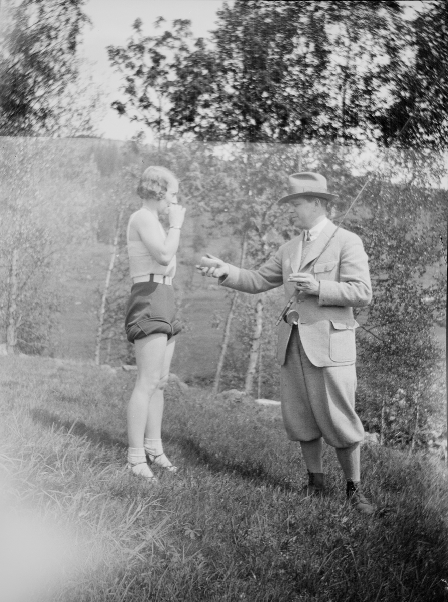En mann kledt i fritidstøy bærer en fiskestang samtidig som han strekker fram hånden og byr en dame, også kledt for fritid, på sigaretter. De står på en røff gressplen kantet med løvtrær.