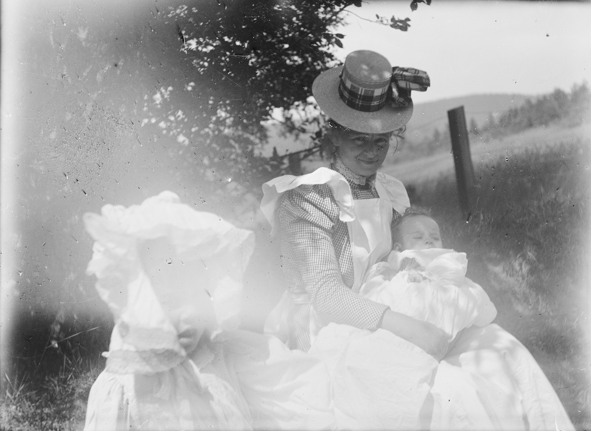 En kvinne sitter i en gressbakke sammen med to barn, et spedbarn og et litt større jentebarn kledt i blonder.