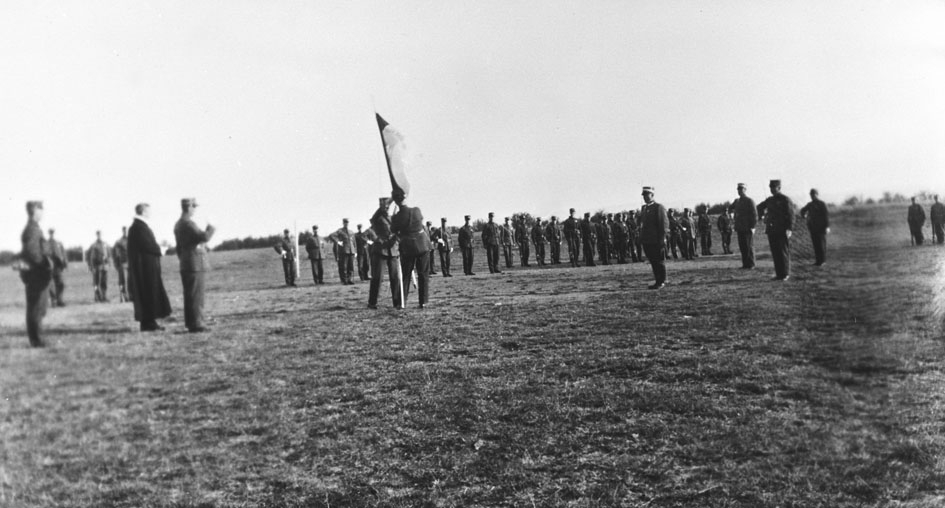 Vi antar at bildene er tatt i forbindelse med gjenopprettelse av Varanger batlajon 01.07.1934. Bildet viser soldater og offiserer, blant dem Prost Skaugen og Genaralmajor Johannesen på eksersersplassen på Nybergmoen 1934.    Et flagg settes i bakken.