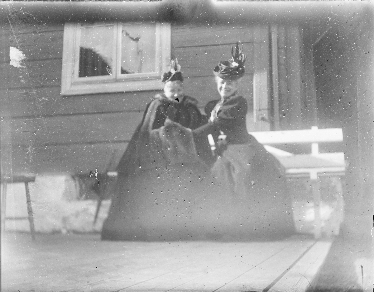 To damer sitter på en benk som står på en platting utenfor et hus.