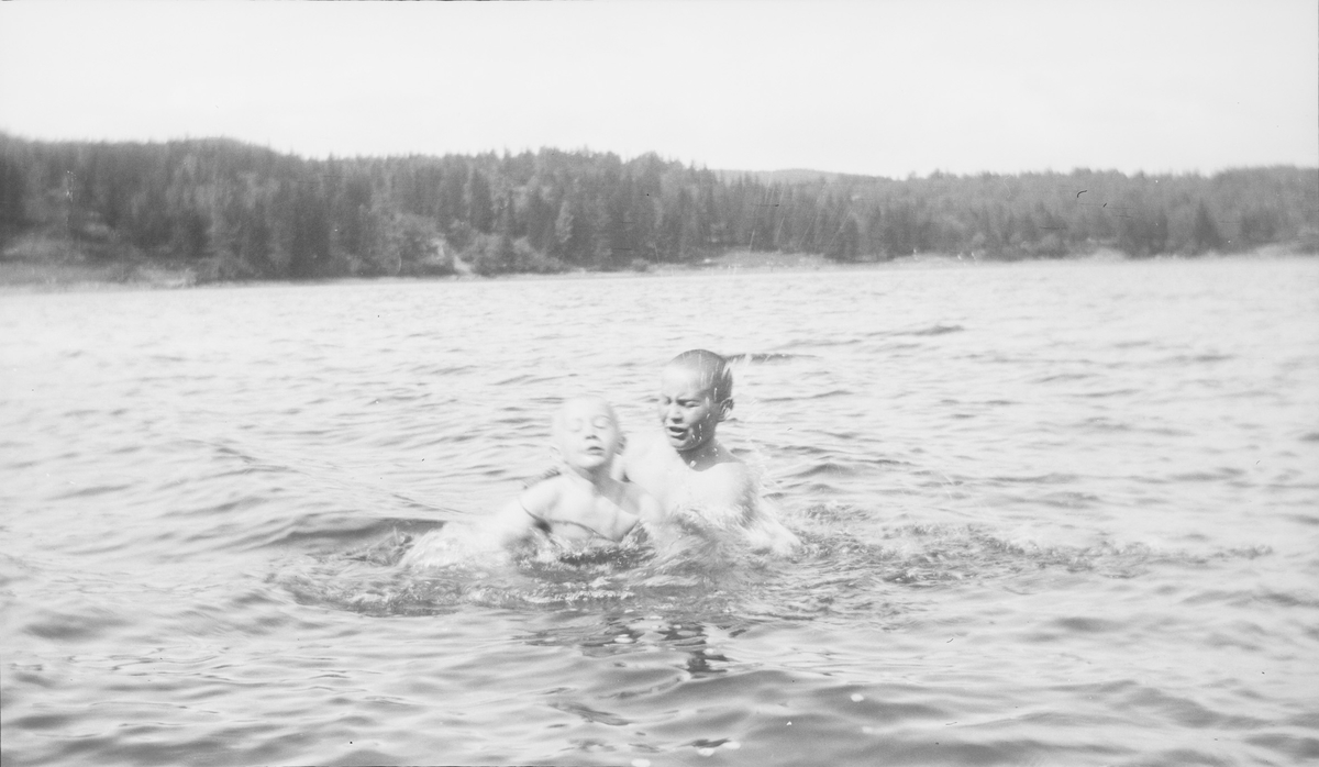 En eldre gutt hjelper en yngre gutt med svømmetrening i en innsjø på østlandet. Den yngste gutten har på svømmebelte. En "kruset" vannoverflate forteller om litt vind. I bakgrunnen sees skog med noen hogstflater.