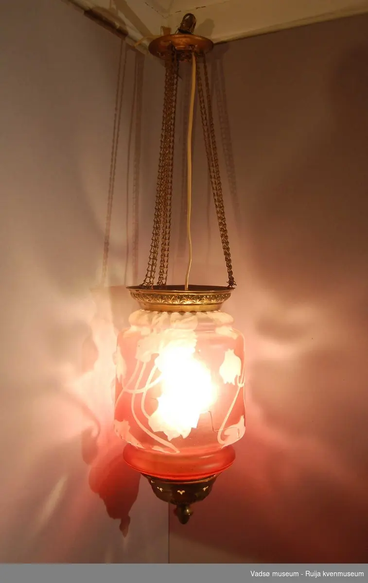 Sylinderformet måneskinnslampe i rosa glass og messing. Glasset har blomsterdekor i relieff. Lampehuset henger i fire messinglenker som går til et sirkulørt takfeste. Lampas øvre kant, bunnstykke og takfestet er i messing.