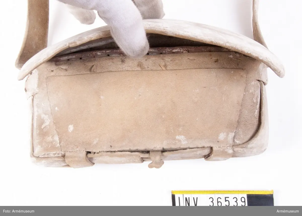 Patronväska av vitt sämskat läder.
Inuti patronväskan finns en bleckplåtsinsats med rum för 12 patroner av ca 18 mm kaliber. Ett par rör saknas och under locket finns en extra ficka.