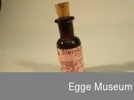 En liten medisinflaske. Brun flaske med kork i toppen, rosa etikett med sort skrift.