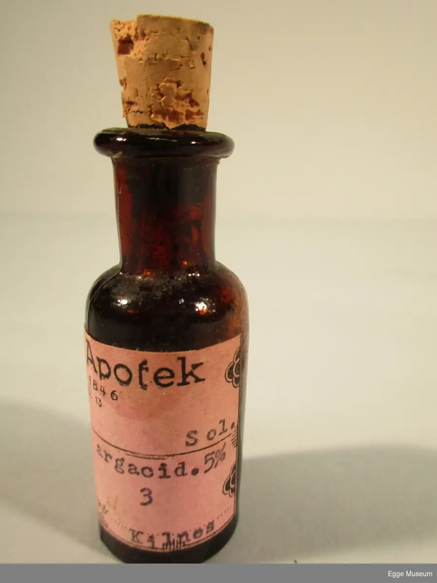 En liten medisinflaske. Brun flaske med kork i toppen, rosa etikett med sort skrift.