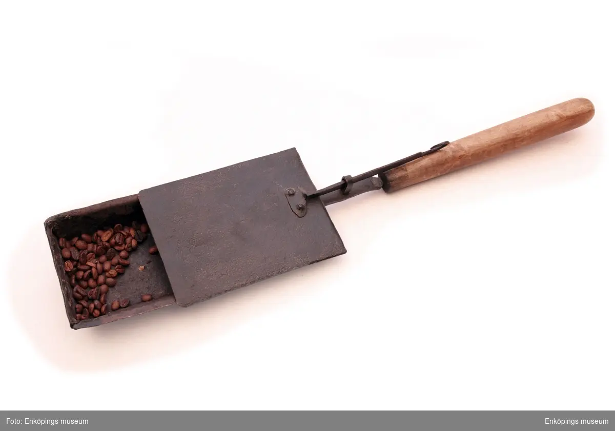 Kaffebrännare av järn med svarvat handtag av trä. Järnlåda med påskjutbart lock 3,5cm hög och 17 x 12,5cm stor. Handtag som är 40cm långt med ett 25cm långt pålagt handtag av trä.