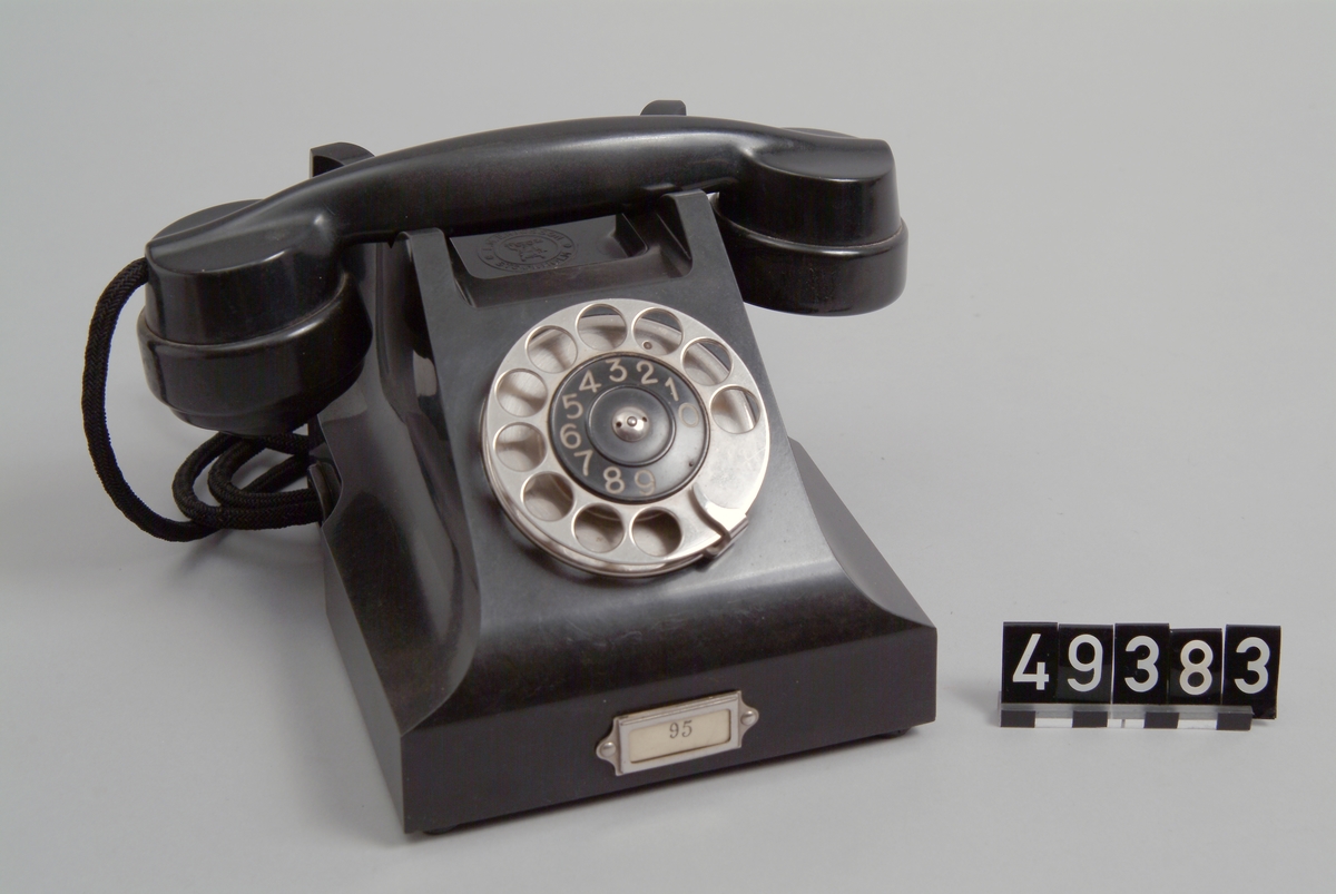 Telefonapparat BC 330, ändstationsapparat för AT-system. Bordapparat modell m33 av svart bakelit med fingerskiva av förnicklad mässing och textilklätt apparatsnöre anslutet till väggplint med lock av svartlackerad plåt.