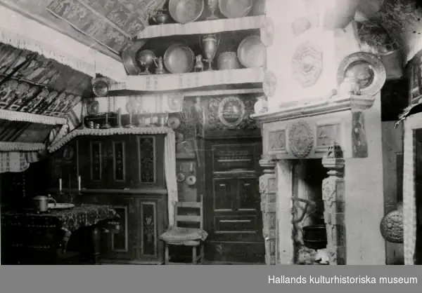Interiör från Bexellska ryggåsstuga, "Bålastugan", då den stod vid Göingegården i Lindberg utanför Varberg (1879-1890). Dragen stuga, med halländska bonadsmålningar och hängkläden. 1600-talsspisen är inte original från Kulsegård i Harlplinge, där stugan stod ursprungligen, utan den installerades vid återuppbyggnaden.
(Se även bild F4217_1)