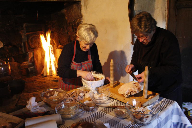Bildet viser to damer som lager sylte til jul. Bak dem brenner gruva, og på bordet foran damene ligger det mange skåler med diverse mat i.