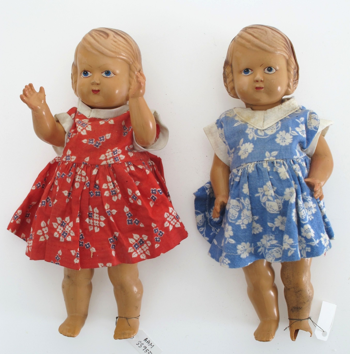 Dukke av støpte plastdeler. Hode og kropp i ett, mens armer og ben er støpt for seg og montert med strikk.   Hjemmesydde klær (?): Rød blomstret kjole. Underkjole. Underbukse i blå blomstet stoff.