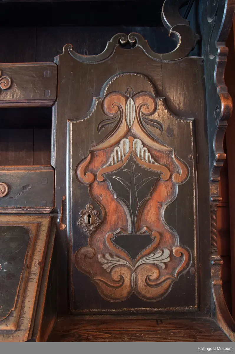 Delene satt sammen v.hj.a. tapping og trenagler.  Gavlen buede sider med skåret kronlist, delt i 3 profilerte, med opprullede ender.  Kronlister fortsetter på sidestykkene.  I toppen av kronlist hjerteformet, gj.brutt utskjæring opp ned.  Gavlens sentralfelt utskjæring i dyp flatskurd.  To løver, vendt mot hverandre, griper om hellebordskaft.  En stake som øverst kløyves i to bjørnehoder i profil skiller løvene.  Gavlen (el. baldakinens) forplate danner fremsiden i skuff i skapets bredde og dybde.  Overskapet:  Åpen hylle øverst, ca. 25 cm høy.  Under hyllen er det delt i 3 felt.  Ett skap i hvert sidefelt.  Midtfeltet har 2 skuffer adskilt av åpen hylle.  H. Skuffe: 12,5 cm.  H. åpent rom: 17 cm.  Under nederste skuff, skap med skråsider som stikker ca. 15 cm ut fra forveggen på overdelen.  Skapet dekkes av liggende skråklaff.  Festet jrenbeslag og hengsler nederst.  
En stor skuff i hele skapets bredde, 6 cm høy nederst, 32 cm dyp.  Over skuffen er rommet delt i 3 felt.  Hvert sidefelt har 2 skuffer og midfeltet en dyp skuff.  Midskuff har fylling med utskåret medaljong, klaff og hylle og skuffen rødmalt. .................................................