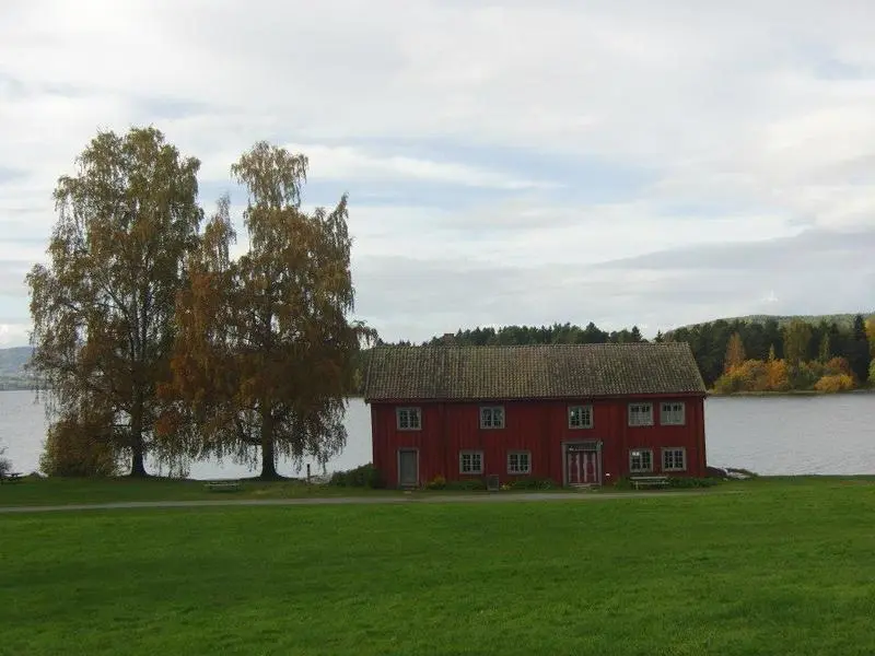 A red, wooden house in two floors are sitting on the banks of the lake with a birch tree to the left.