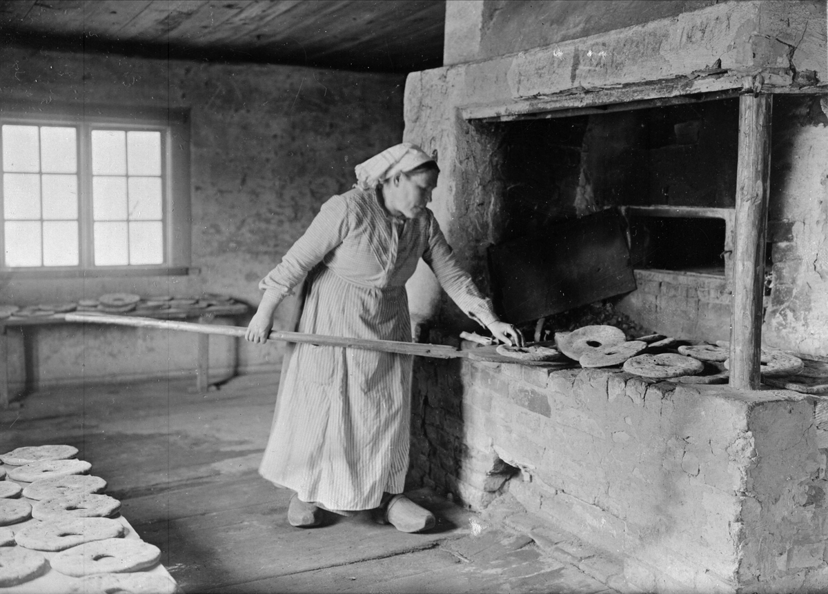 Bakning. Kvinna iklädd huckle arbetar med lång brödspade vid vedeldad ugn. Västergötland.  