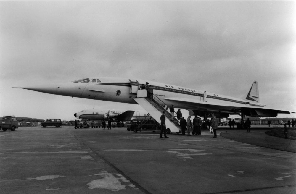 Lufthavn/Flyplass. Bodø. Et fly, Concorde fra Air France, parkert, og stor aktivitet rundt flyet. Ved siden av står et propellfly fra Delta Air Transport parkert.