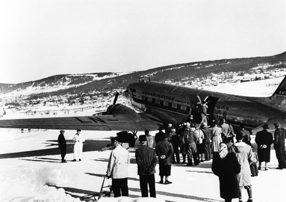 Lufthavn/Flyplass. Et fly, DC-3 fra SAS, parkert i vinterlige omgivelser på Mjøsisen. Det er stor samling av passasjerer som er klare til å gå ombord i flyet.
