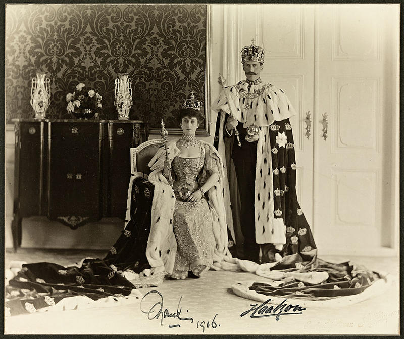 Dronning Maud (1869-1938) og Kong Haakon VII (1872-1957) fotografert i 1906 i Stiftsgården i Trondheim. Begge med kronregalier, dronningen sittende med kappen liggende utover gulvet, kongen stående i tilsvarende kappe.