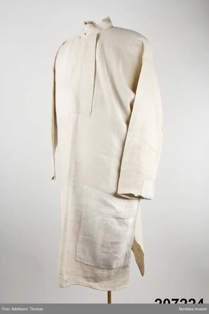 Långskjorta av vit tämligen grov och glesvävd linnelärft.  Ett bålstycke med sidsömmar och sidsprund, vidsydd rak ärm med ärmspjäll.  4 cm hög ståndkrage,  29 cm halssprund med  9,5 cm brett förslag knäppt uppe vid halsen med en trådknapp. Skjortan handsydd. 
Anm. Sliten med några hål i nederkanten, dessutom  lappad med linnetyg  i knähöjd på framsidan, samt på ärmarna, ena ärmen lappad med bomullslärft. 
 Har brukats av hammarsmeden  Janne Valström f. 1852,  vid  Strömbacka bruk i Hälsingland. Smederna bar linneskjorta, träskor och benholkar som skydd mot värmen.
/Berit Eldvik 2012-12-20