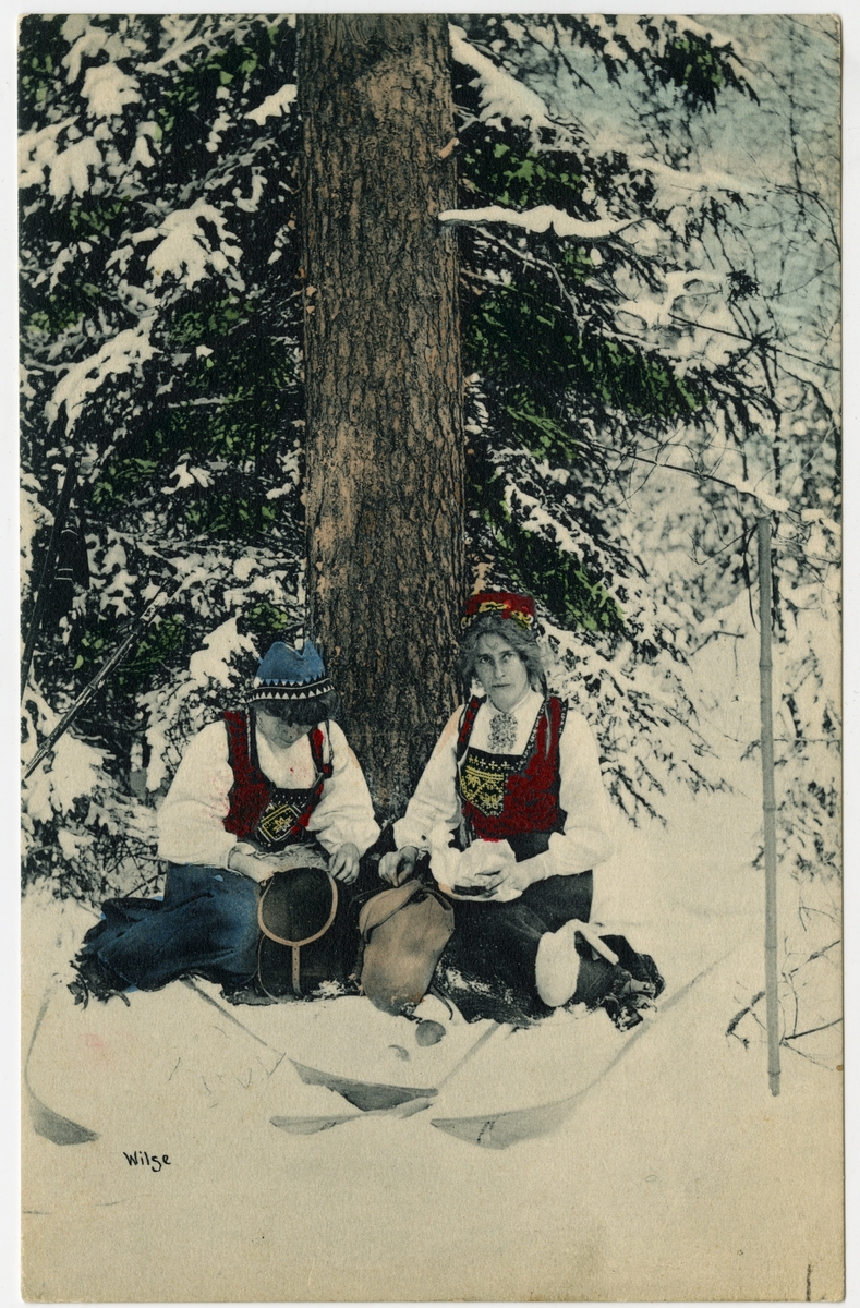 Kolorert postkort. To kvinner i bunad sitter i snøen under et tre.