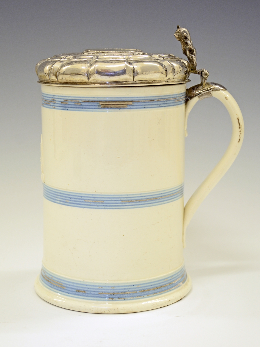 Fra protokollen: 
Steintykrus med sylvlok. Kruset sjølv cylindriskt med 3 blaa, rundu belte paa den kvite glasuren. Framme eit upphøgja felt i eggform med eit tverband yvi med stempla: CR-IV IMPERIAL.
Loket med støypt ciselera løve (med øks i labbane) til lokknapp og uppdriven runding i midten med innfelt sylvpening (medalje merkt F.3, 1659, arm fraa sky høgg av hond som grip etter ei krune). Voluten ikringum bytt med knekkir, og flaten rundt peningen med uppdrivne ciselera granne bandknekkmynster paa mattera grunn. Regence-stil; truleg arbeid av ein bygdesylvsmed (utan stempel). Paa volutknekkine er gravi einstad J. V. B., einstad M. H. D. H. og 1735. Høgd med loket 18,5 cm, tvermaal paa loket 11 cm. Stemp: CM.