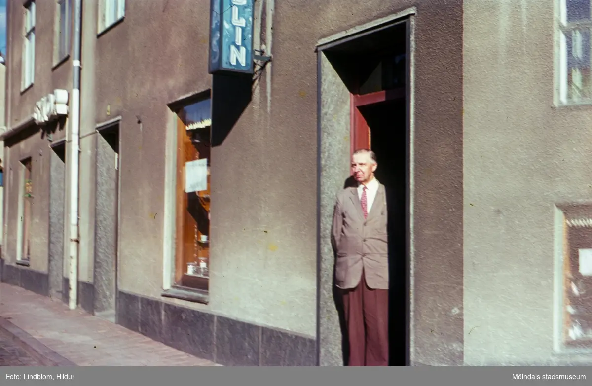 En man står i dörren till Josef Anderssons Porslinsaffär med adress Kvarnbygatan 4 vid Gamla Torget i Mölndal, 1960-1970-tal. I samma hus låg även Rasmussons skoaffär.

För mer information om bilden se under tilläggsinformation.