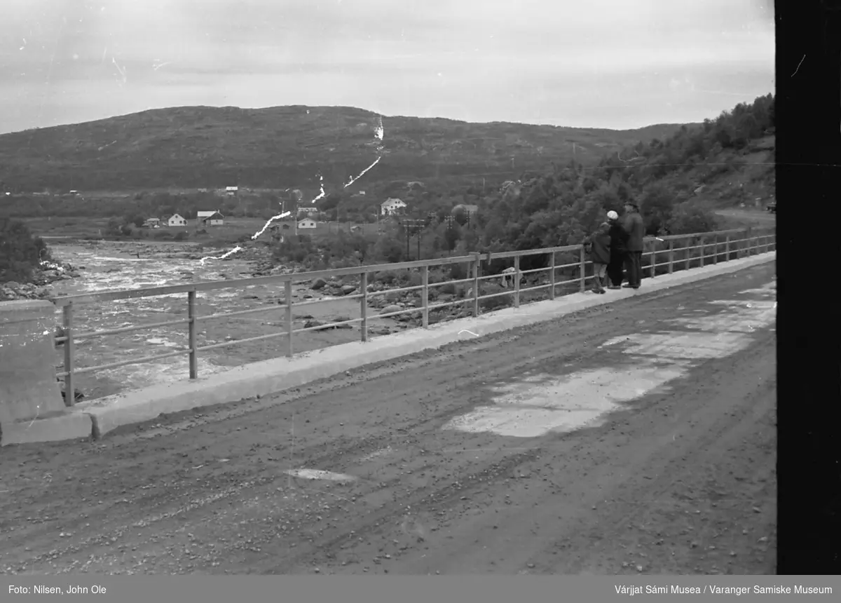 Tre personer står på Neiden bru og ser på elva. Bebyggelse i bakgrunnen og et telt nede ved elvebredden. 1967.