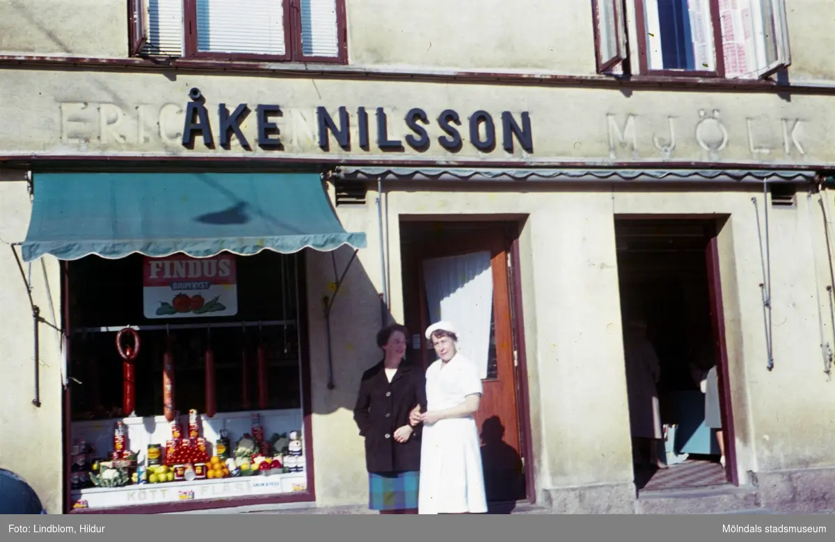 Två kvinnor står utanför dörren till Åke Nilsson charkuteri och delikatesser med adress Kvarnbygatan 43 vid Gamla Torget i Mölndal, 1960-1970-tal.

För mer information om bilden se under tilläggsinformation.