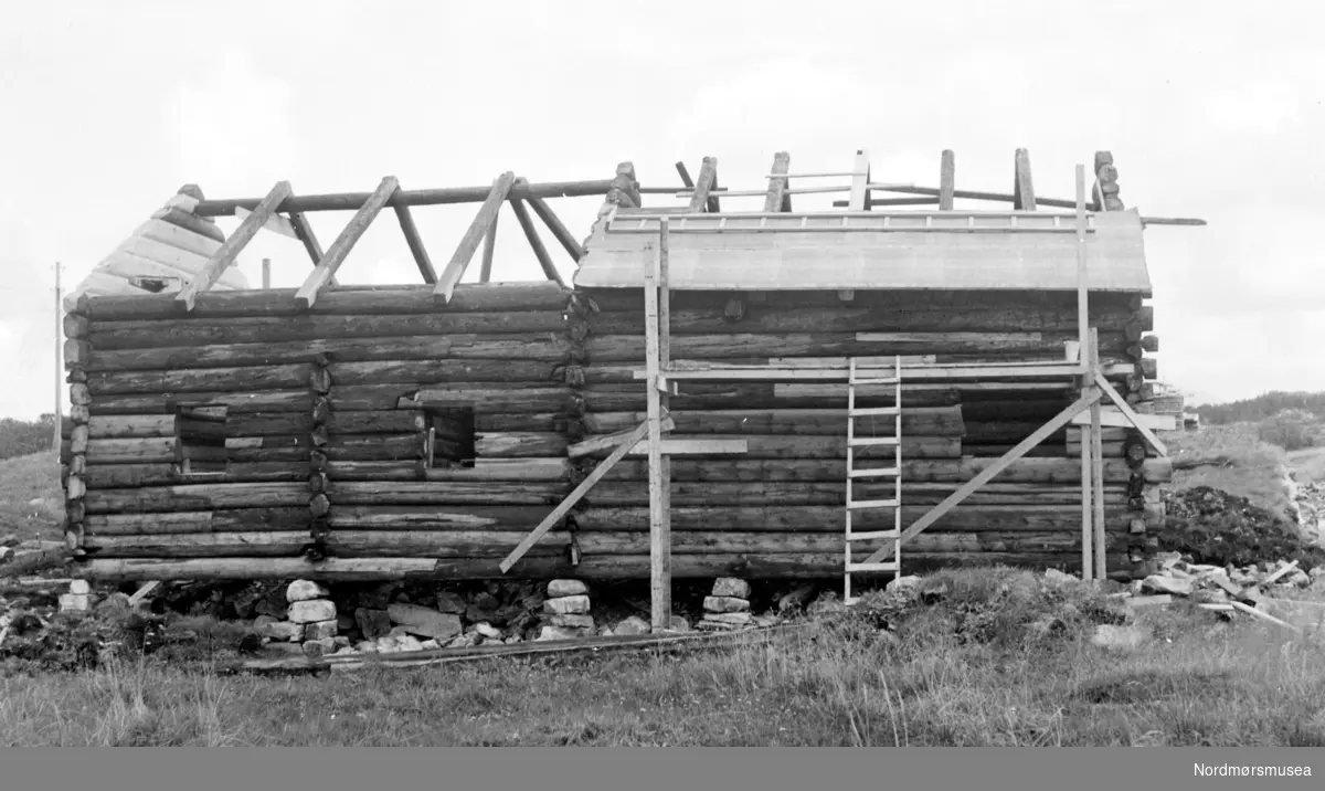 På bildet ser vi Almskårstua under oppføring i Knudtzondalen, etter at den har blitt flyttet fra Clausenengen. Her skal taket legges, og vi ser takkpappen tilhøyre. Bildet er fotografert fra vest, og datert 25. august 1959. Nordmøre Museum.