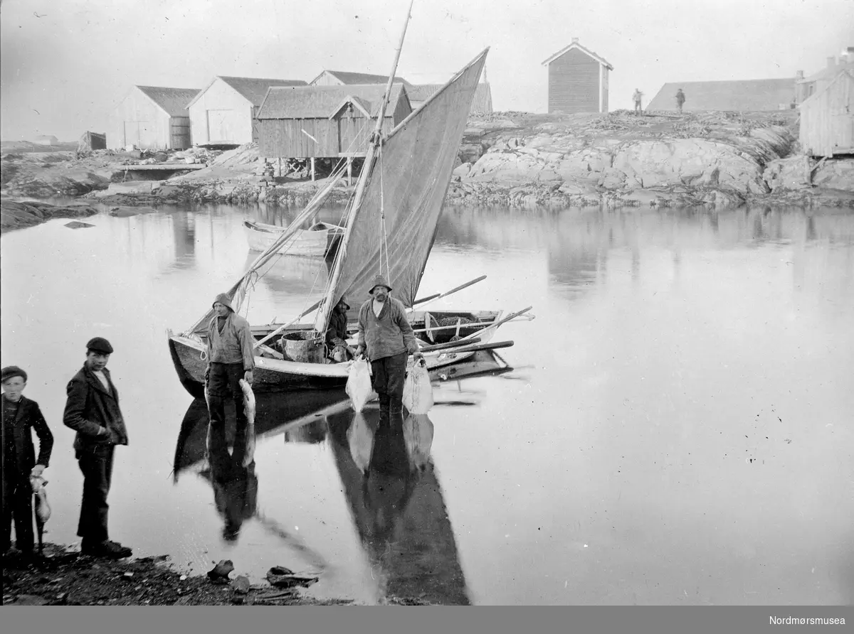 Foto trolig fra
Breivågen, muligens fra Veiholmen i Smøla kommune, hvor vi ser to fiskere med sin seilbåt. Legg merke til de to guttene foran i ytterkanten på venstre side. Fra Nordmøre museums fotosamlinger.
