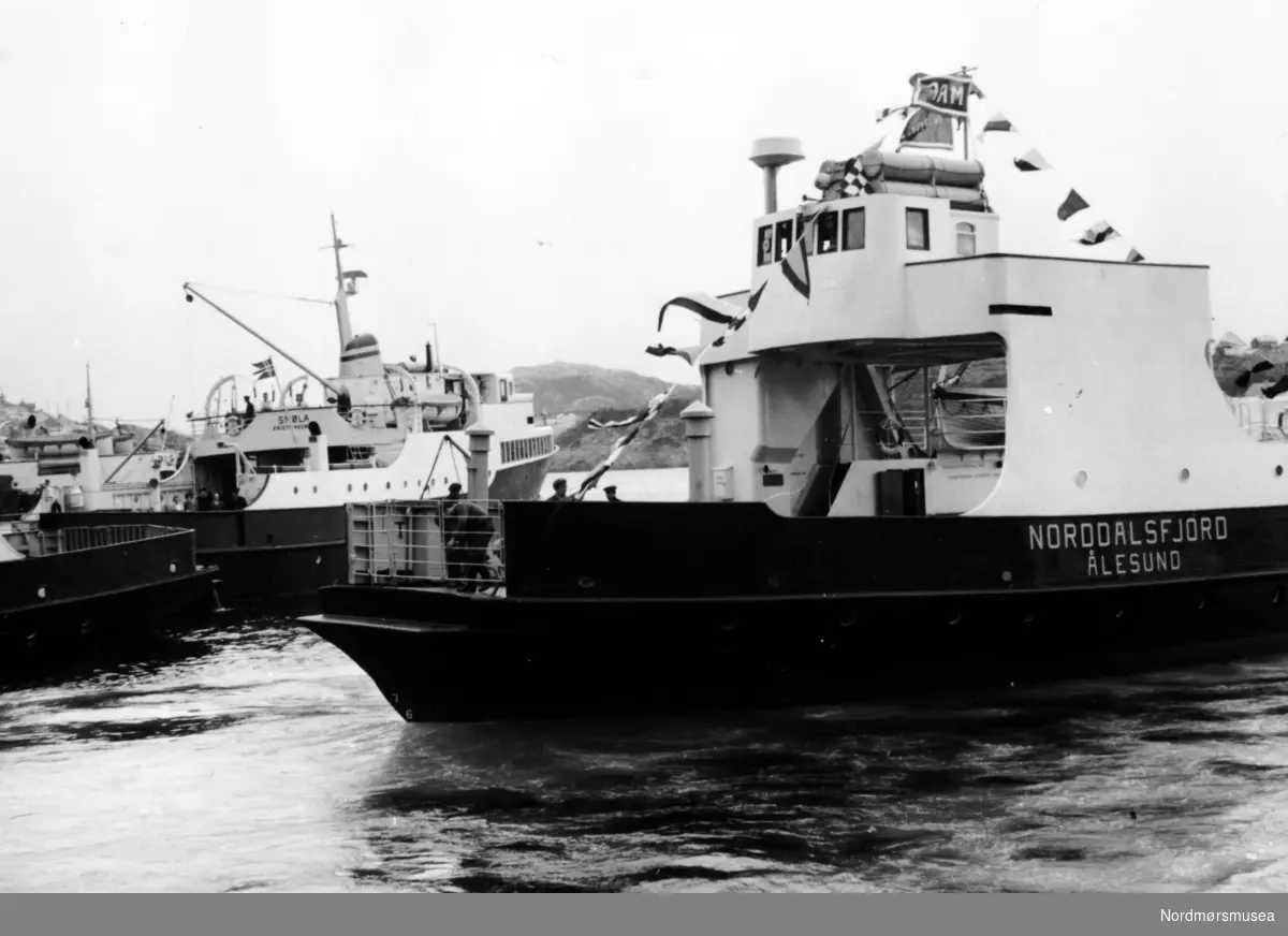 Bildet viser B/F"Norddalsfjord" Storviks Mek. Verksteds bnr.14 som legger fra kai i huken ved fergetillegget på Nordmørskaia etter overleveringsturen. Vi ser at signalflaggene og rederiflagget er heist. 

"Norddalsfjord" ble levert til Møre og Romsdal Fylkesbåtar 15. mars 1961 og hadde følgende hoveddimensjoner: L 31,20 m x B 8,55 m x D 3,35 m og hadde en tonnasje på 159 bruttoregistertonn. Fremdriftsmaskineriet består av 3 Volvo Penta turboladede dieselmotorer type TMD96 på til sammen 420 hk som via kilremdrift var koblet til et felles gir og propellaksel med vribar propell, slik at hver enkelt av motorene kunne kjøres separat. Fergen hadde 2 Bolinders vekselstrømsaggregater type 1052MG på 23 hk hver tilkoblet en generator på 17 kW. Fergen var utstyrt med elektrohydraulisk styremaskin.

Fergen har plass til 18 personbiler og har sertifikat for 160 passasjerer. Forut er det innredet 6 lugarer for offiserer og restaurantpersonale og akterut en mannskapslugar for 4 personer og toppfarten er 11,4 knop og marsjfarten 10,5 knop.

Ferga er verkstedets første nybygg etter B/F"Trygge" som ble levert i 1938.

Personene, på dekket, fra verkstedet er vanskelige å kjenne igjen.

I bakgrunnen ses B/F"Smøla" og 2 andre ferger. Fergen bak Smøla kan være B/F"Geiranger" (B/F"Trygge" ?).

Bildet er fra 1961. Kilde/tekst: Peter Storvik. Fra Nordmøre museums fotosamlinger.
