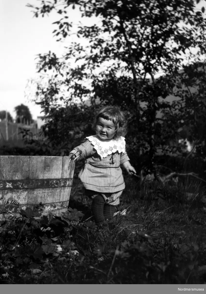 Portrett av et barn som står ved en trestamp i en hage. Fra Nordmøre museums fotosamlinger.