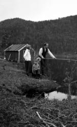 En mann fisker. En kvinne og to barn ser på. Vi ser et naust