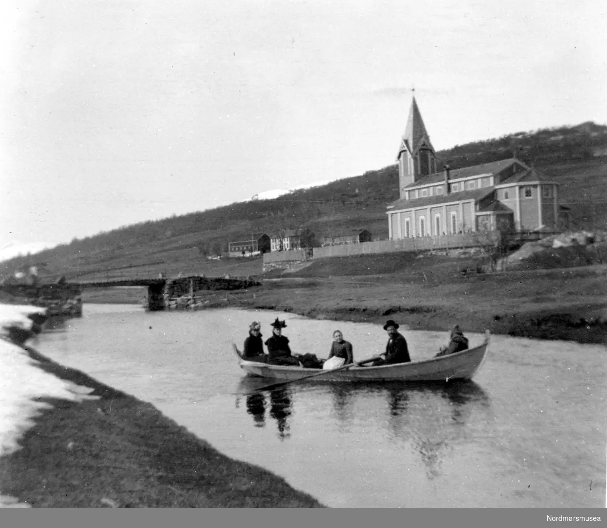 Foto fra Nesset kirke i Eidsvåg, Nesset kommune, hvor vi ser en en liten gruppe kvinner og en mann ute i en robåt på den vesle elven. Fotograf er Georg Sverdrup. Datering er mer usikker, men trolig fra perioden 1930 til 1939. Fra Nordmøre Museums fotosamlinger.
