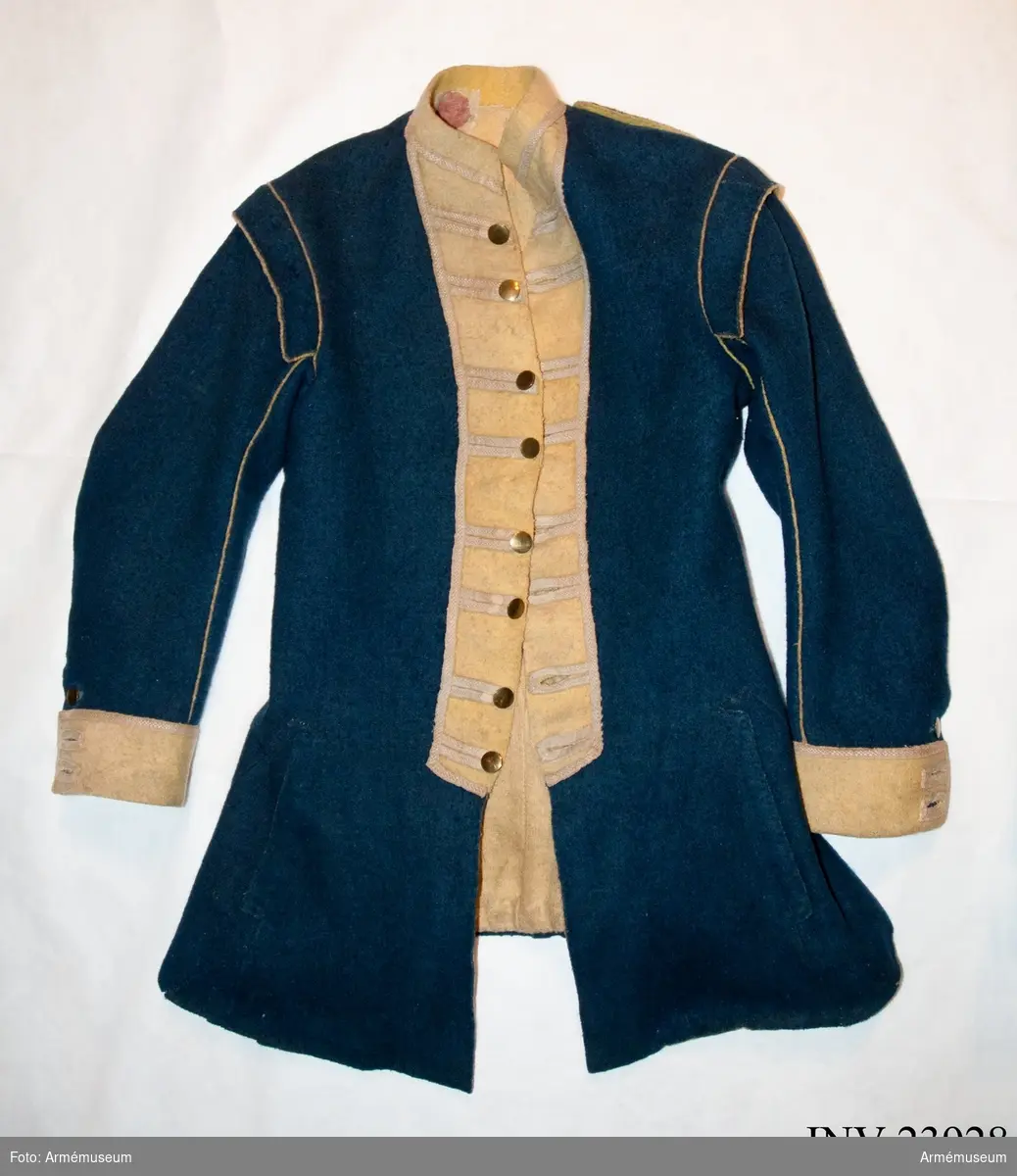 Jacka av blått kläde med gula bröstrevärer, krage och ärmuppslag, fodrad med gul boj samt linne i ärmarna. Försedd med mässingsknappar och vävda band som dekoration.