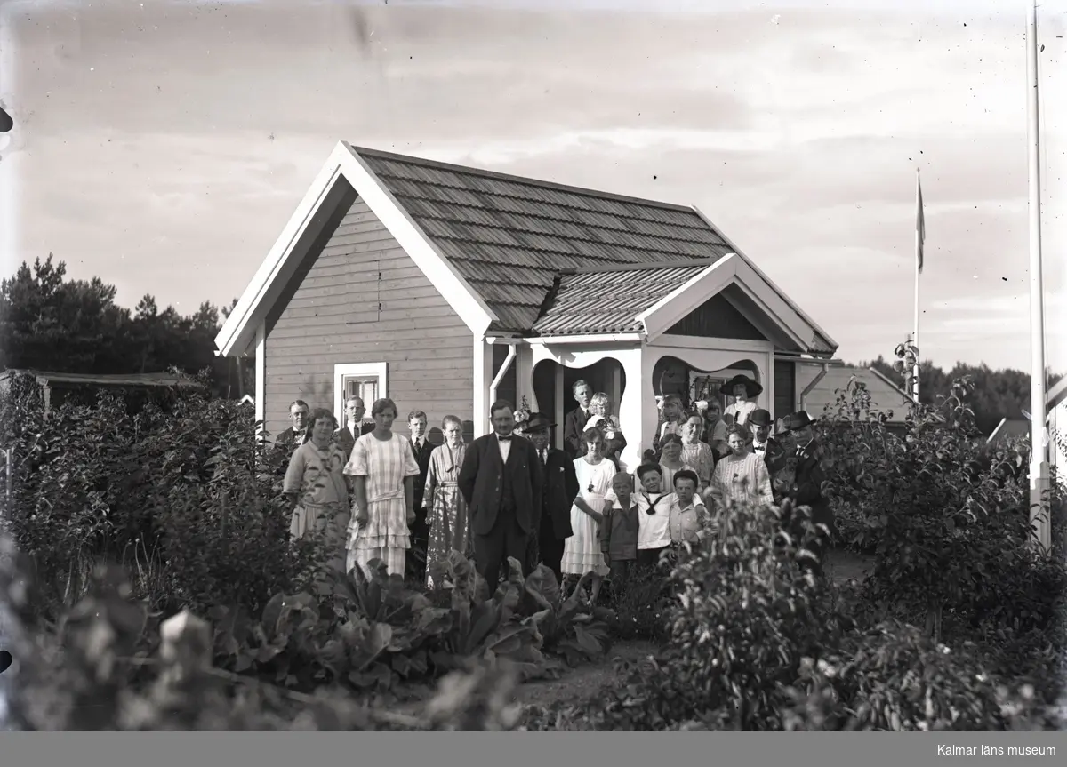 En stuga och en grupp människor i Kalmar södra koloniområde, fotograferat omkring 1930. Kalmar södra koloniförening grundades 1917 och har idag 105 kolonilotter. Området ligger strax söder om länssjukhuset i Kalmar med huvudingång från Stensbergsvägen.