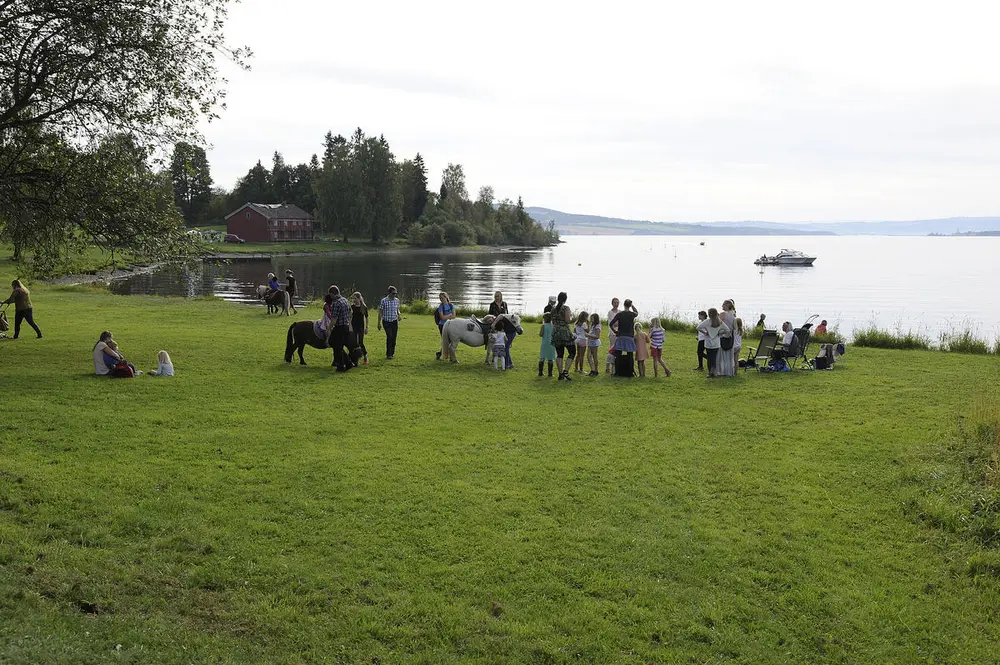 Barn og ungdom som driver med aktivitet på grønn gresslette ved vannet, museumshus i bakgrunnen.