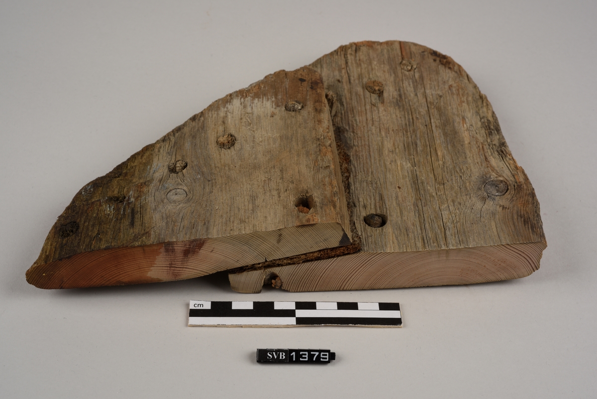 Del av et båtbord. Delen ble saget av. Det er hull til vidje og plugger. Delvis er det rester av både vidje og plugger. Treet fremstår ubehandlet. Det er et materiale som ligner torv/mose mellom bordene.
