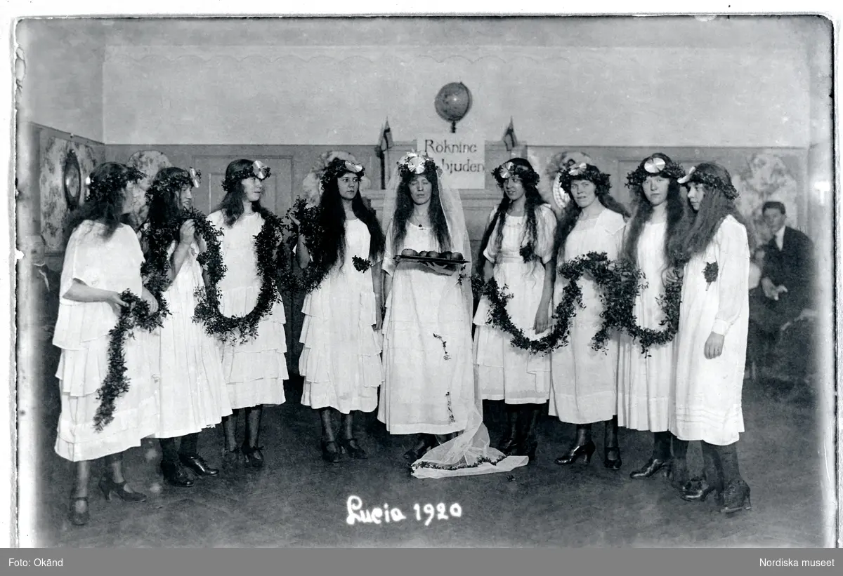 Luciafest i gamla Tranebergsstugan 1920. En grupp unga kvinnor är klädda i vita särkar och bär kransar av lingonris som huvudprydnad. I mitten står Lucia med en bricka, tärnorna håller en lång girland av lingonris.