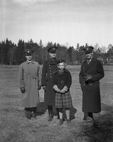 Tre uniformerade herrar (varav en polis ?) och en flicka med skolmössa står på en äng. I bakgrunden skymtar en militärbarack (?).