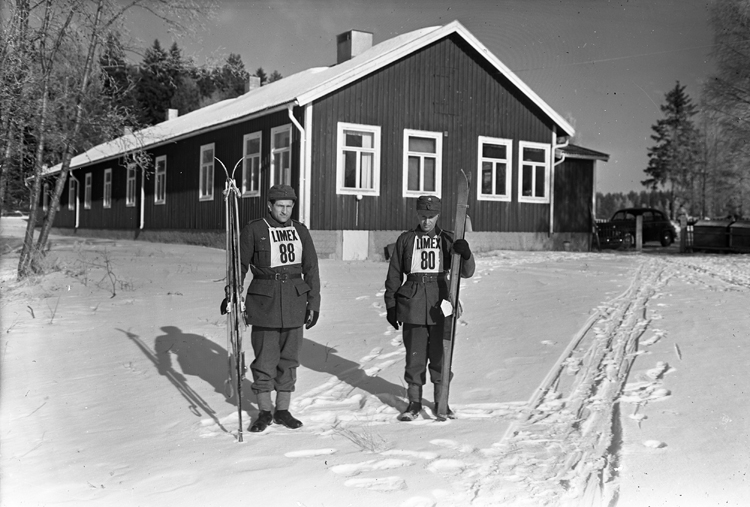 Två skidlöpare står framför en barack en vinterdag.
I bakgrunden syns en bil.
"Limex" - skidfabriken Limex i Tobo bruk, Tegelsmora socken,
Uppland.