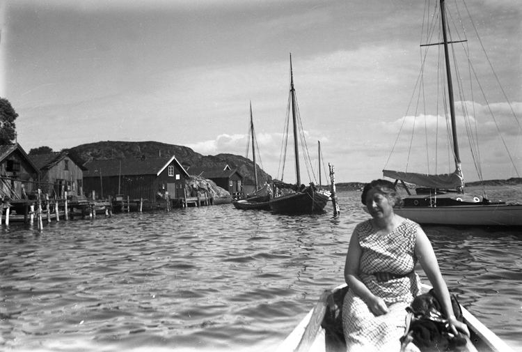 En kvinna sitter i en båt.Bakom henne syns en hamn med några bodar och brygga.