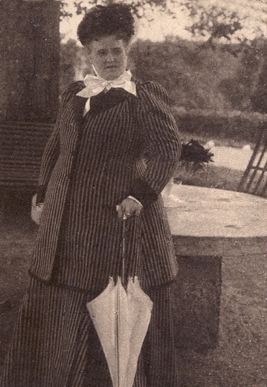 Portätt av Christina Nilsson  där hon står i en park. Hon bär kavaj och kjol samt någon form av huvudbonad. I handen håller hon ett paraply.

Möjligen vid Gårdsby säteri (?) (AB).