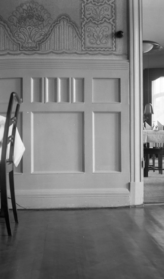 En vägg, där övre halvan är tapetserad och undre halvan utsmyckad i trä. I förgrunden kan en stol skymtas och i bakgrunden stolar och bord genom en dörröppning.

Interiör från matsal på Industri-restaurangen i Kristianstad 1943